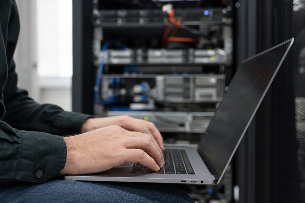 Administrator typing on a laptop while reviewing server patching tasks, with a blurred server rack behind.